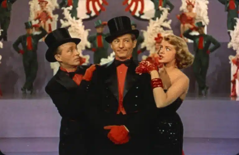 Bing Crosby, Danny Kaye, and Rosemary Clooney in formal black attire and top hats stand on stage, surrounded by dancers in red and green costumes. The scene feels festive and lively.