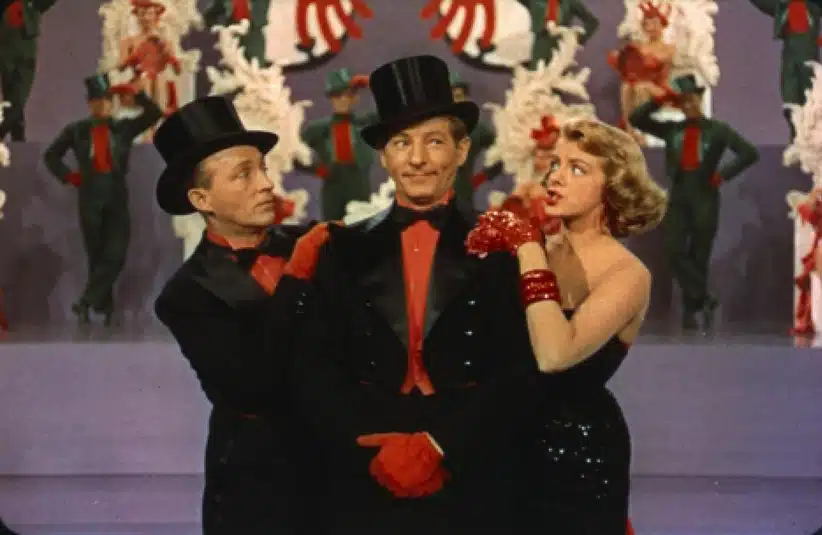 Bing Crosby, Danny Kaye, and Rosemary Clooney in formal black attire and top hats stand on stage, surrounded by dancers in red and green costumes. The scene feels festive and lively.