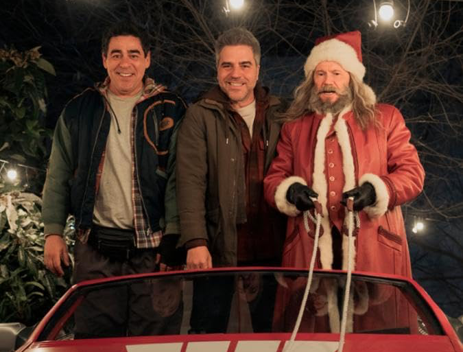 Three men stand outdoors at night, smiling warmly. One is dressed as Santa Claus, holding a sleigh rope. Festive lights and trees frame the scene.