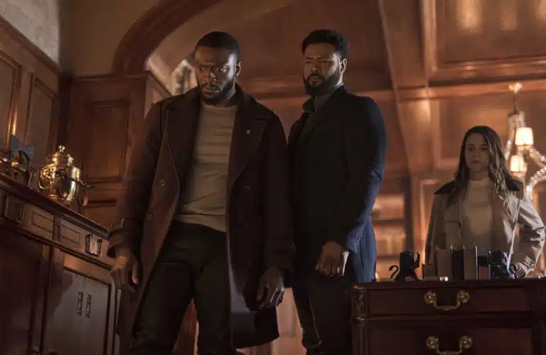 Aldis Hodge, Alona Tal, and Isaiah Mustafa in "Cross (2024)" stand pensively in an elegant, wood-paneled room with ornate decor. The atmosphere is tense, hinting at a serious or dramatic moment.