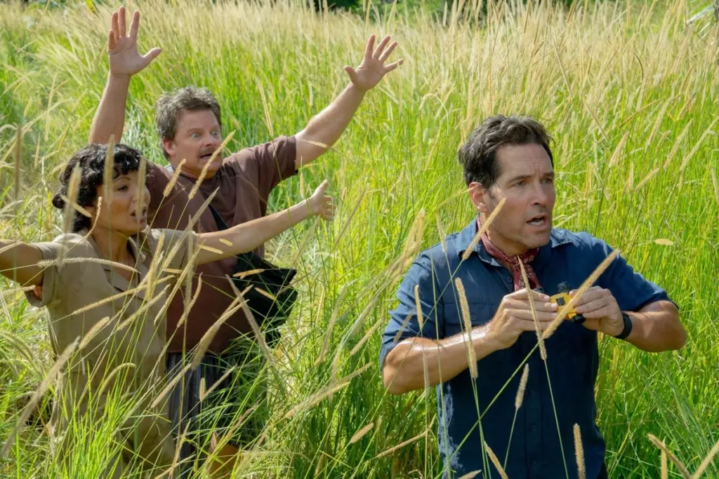 Three people stand in tall grass, looking surprised and excited. The person in front holds a small object, while the others have arms raised in enthusiasm.