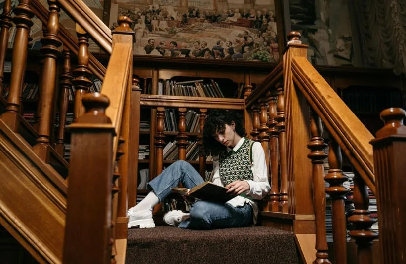 A student finding a reading nook on a staircase landing of a library, reading in pease, cozy, winter time