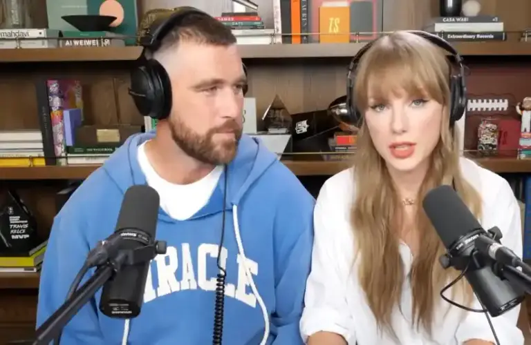 Travis Kelce in a blue hoodie and Taylor Swift in a white shirt, both wearing headphones, speak into microphones on Image of Taylor Swift and Travis Kelce on the New Heights podcast via NHTV Youtube with a bookshelf with books and memorabilia is behind them.