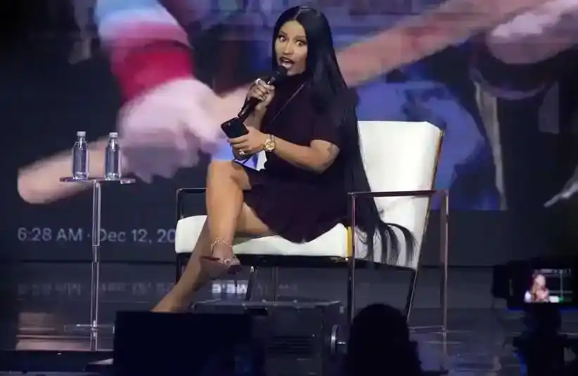 Nicki Minaj with long black hair sits on stage in a white chair, holding a microphone at Turning Point USA's AmericaFest. She is dressed in black and smiling, she appears confident and engaged.
