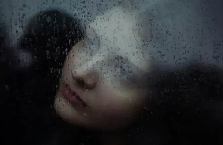 Woman looking through a window covered in raindrops, loneliness