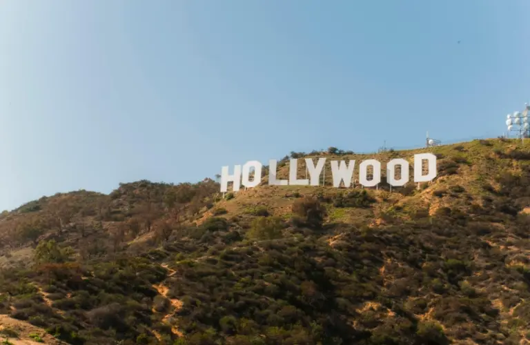 The iconic Hollywood sign is set against a clear blue sky atop a sunlit, grassy hill. It conveys a sense of fame and the film industry's allure.