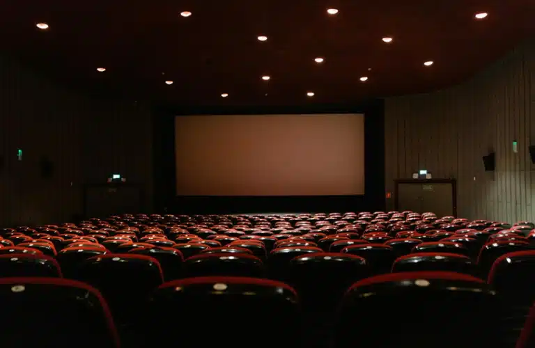Empty movie theater with rows of red seats facing a large blank screen. Soft ceiling lights create a calm, expectant atmosphere.