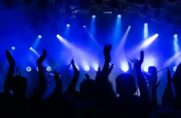 Silhouetted crowd with raised hands at a concert, illuminated by vibrant blue stage lights, creating an energetic and lively atmosphere as music from a 2025 pop album plays.