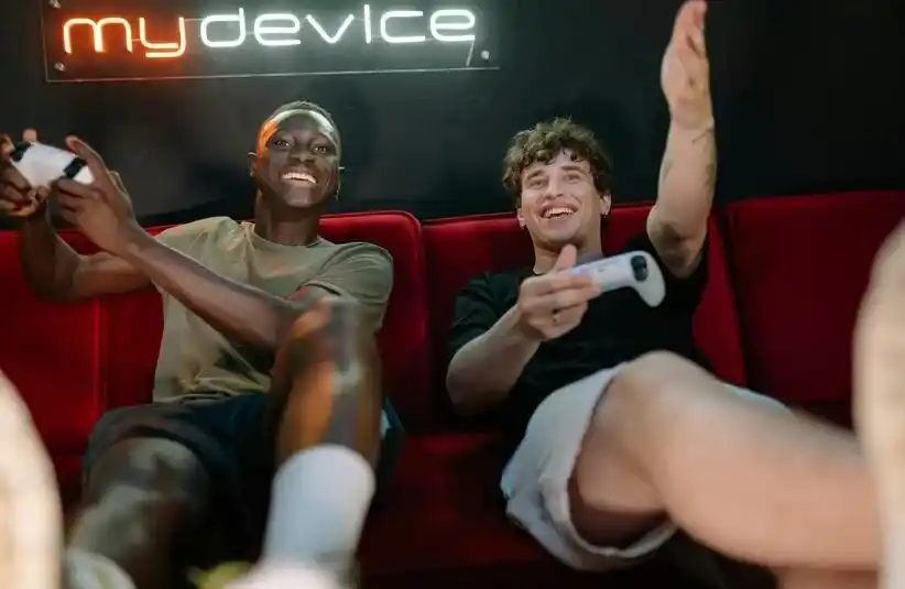 Two friends are joyfully playing video games on a red couch, holding controllers. A neon "mydevice" sign glows in the background, creating a fun, relaxed atmosphere.