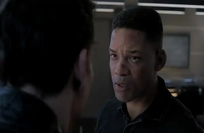 Will Smith looks intently at another person in a dimly lit room, appearing emotional and serious. The background is blurred, emphasizing the intense exchange.