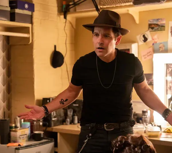 Paul Rudd in a black t-shirt and hat stands in a cluttered dressing room, looking puzzled and unstoppable, holding a red lollipop. Bright lights, casual atmosphere.