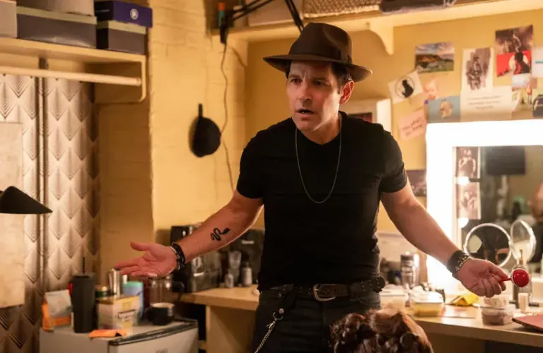 Paul Rudd in a black t-shirt and hat stands in a cluttered dressing room, looking puzzled and unstoppable, holding a red lollipop. Bright lights, casual atmosphere.