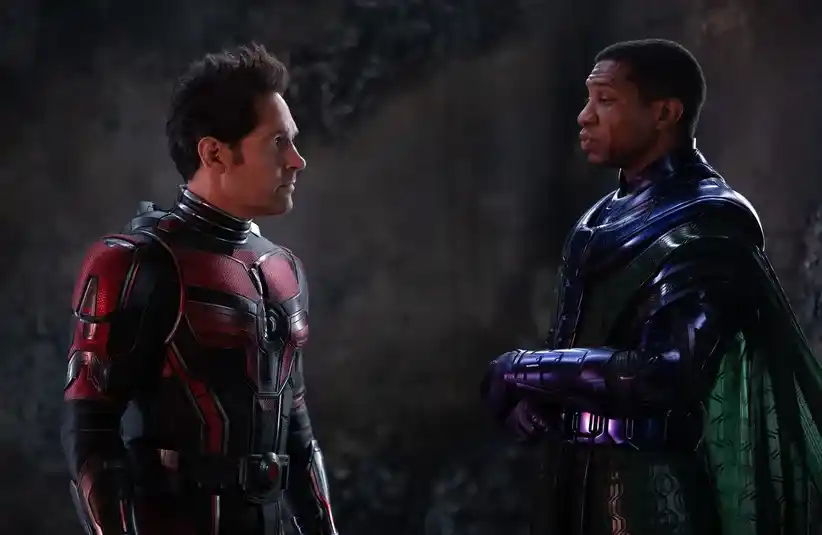 Paul Rudd and Jonathan Majors in Ant-Man and the Wasp, standing in a dim setting, engaged in a tense conversation. The left one wears a red and black suit, while the right wears dark armor with a cape.
