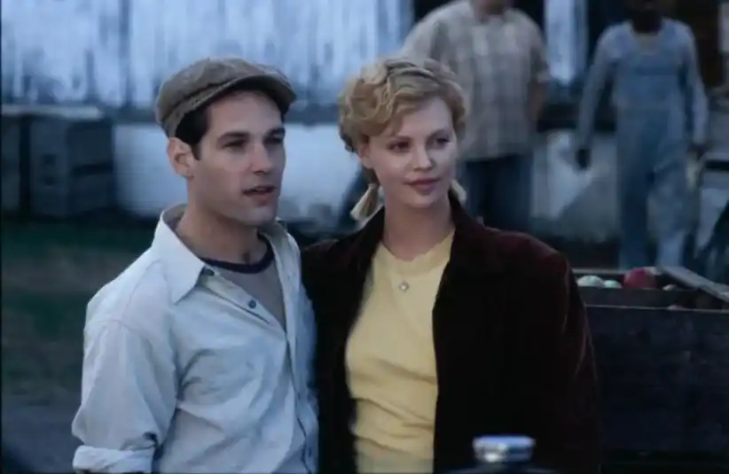 Paul Rudd and Charlize Theron stand close, smiling, in a rustic outdoor setting, in "The Cider House Rules." The man wears a cap and shirt, while the woman has short curly hair and a jacket. Slightly blurred people and crates are in the background, creating a warm, nostalgic mood.