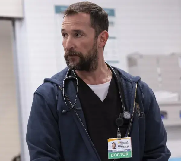 Golden Globes winner, Noah Wyle in The Pitt (2025), wearing a stethoscope and ID badge labeled "DOCTOR," stands in a hospital setting. He has a thoughtful expression, conveying professionalism.