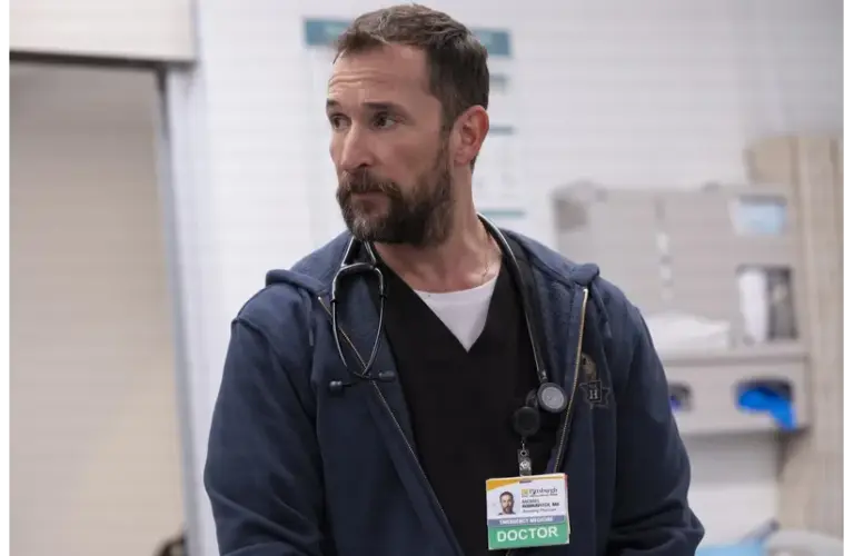 Golden Globes winner, Noah Wyle in The Pitt (2025), wearing a stethoscope and ID badge labeled "DOCTOR," stands in a hospital setting. He has a thoughtful expression, conveying professionalism.