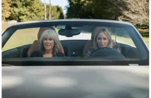 Catherine O'Hara and Emily Blunt sit in a convertible car, driving down a country road surrounded by greenery. They appear relaxed and content, enjoying the sunny day.