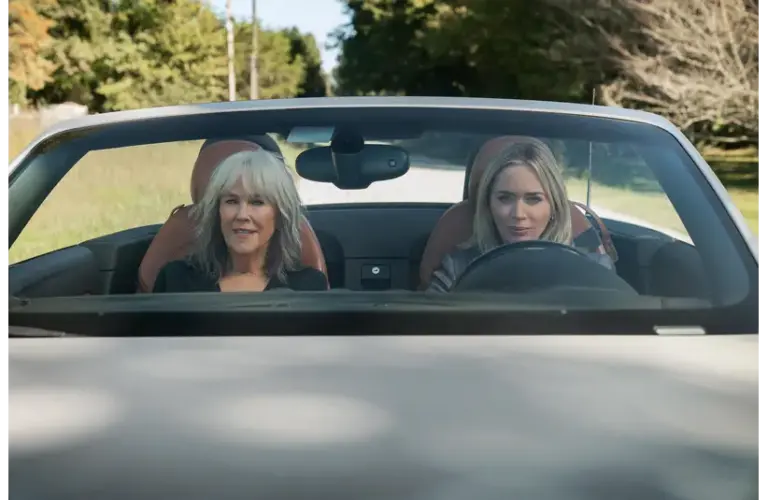Catherine O'Hara and Emily Blunt sit in a convertible car, driving down a country road surrounded by greenery. They appear relaxed and content, enjoying the sunny day.
