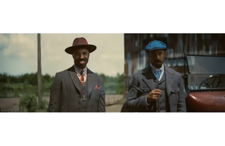 Michael B. Jordan in Golden Globes-winning "Sinners (2025)" as Smoke & Stack in vintage suits stand outdoors near a classic car. The man on the left wears a burgundy hat, while the right wears a blue cap. The scene feels nostalgic.