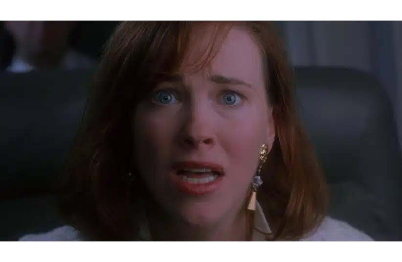 Catherine O'Hara, a woman with red hair looks shocked and worried, her mouth open and eyes wide. She wears gold earrings and sits in a dimly lit setting, conveying tension. She is about to say "KEVIN!" in Home Alone.
