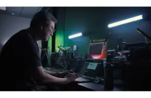 An NVIDIA employee works intently on a laptop in a dimly lit tech lab filled with equipment for the NVIDIA G-SYNC. Two fluorescent lights cast a focused, industrious atmosphere.