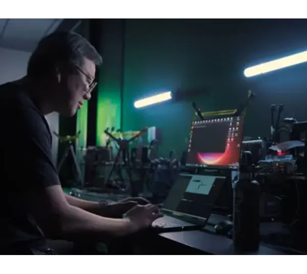An NVIDIA employee works intently on a laptop in a dimly lit tech lab filled with equipment for the NVIDIA G-SYNC. Two fluorescent lights cast a focused, industrious atmosphere.