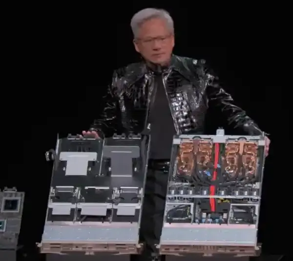 NVIDIA CEO Jensen Huang speaking live at CES 2026, he is in a shiny black jacket presents two large computer hardware components on stage. The components are detailed, with visible wires and circuits.