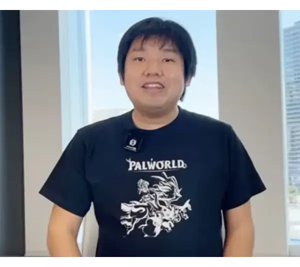 Poketpair CEO, Takuro Microbe, in a Palworld t-shirt, smiles in a bright office with urban buildings visible through large windows. The atmosphere is casual and friendly.