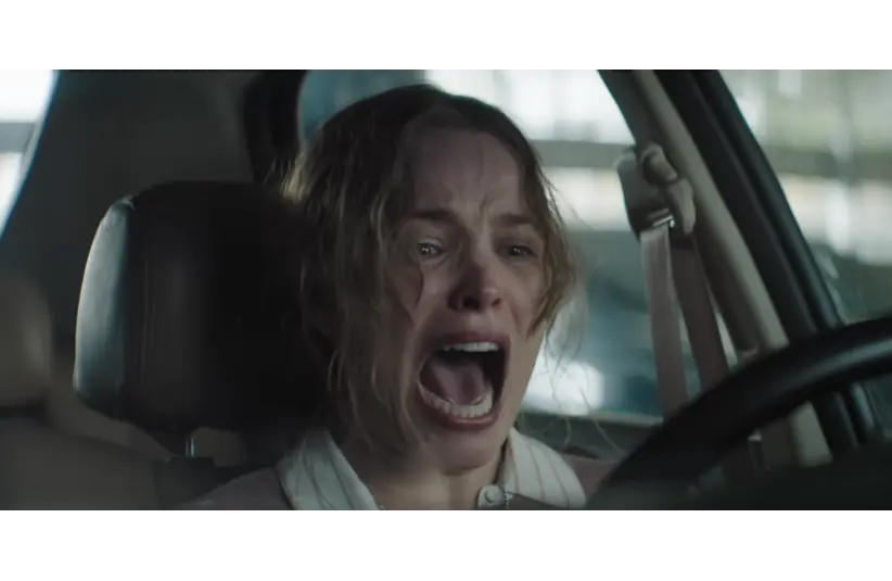 In "Send Help, "a a woman with wavy hair is seated in a car, gripping the steering wheel, her mouth open in a scream. Her expression conveys fear and panic.