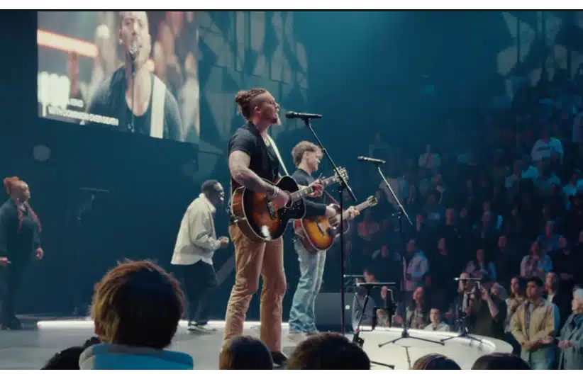 Brandon Lake and Elevation Worship perform on a stage in front of a large audience, with a screen showing a close-up of a singer. The atmosphere is vibrant and dynamic.