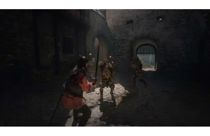 A medieval battle scene shows a warrior in red confronting two armored knights with swords in a shadowy stone courtyard, conveying tension and bravery. The Blood of Dawnwalker, Courtesy of Rebel Wolves and Bandai Namco Entertainment.