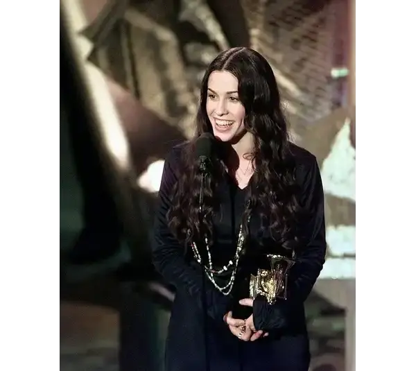 Alanis Morissette is a 2026 inductee for the Songwriters Hall of Fame. She has long dark hair, smiles joyfully while holding a trophy in one hand and speaking at a microphone, set against an abstract background.