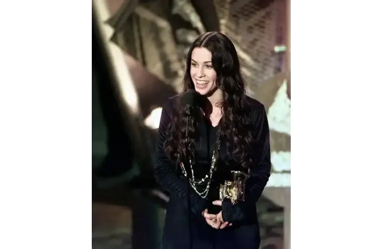 Alanis Morissette is a 2026 inductee for the Songwriters Hall of Fame. She has long dark hair, smiles joyfully while holding a trophy in one hand and speaking at a microphone, set against an abstract background.