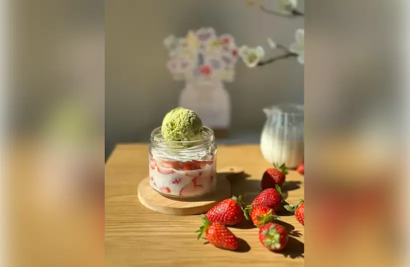 A cup of strawberries and cream with a scoop of vibrant green matcha sorbet on top. Fresh strawberries are scattered eloquently on the table beside the cup