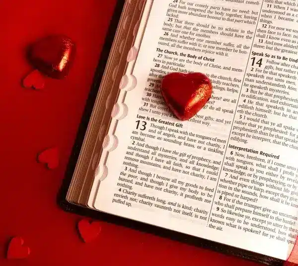Open Bible on a red surface, displaying the "Love is the Greatest Gift" passage, surrounded by shiny heart-shaped candies, conveying warmth and love., Saint Valentine, Valentine's Day