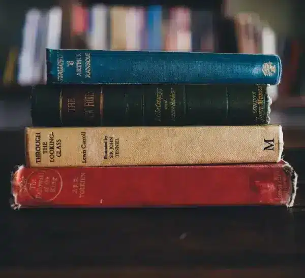 A stack of four vintage books with worn covers sits on a dark wooden table, set against a blurred background of a bookshelf, evoking a nostalgic library feel. Could they be part of the International Booker Prize list of 2026?