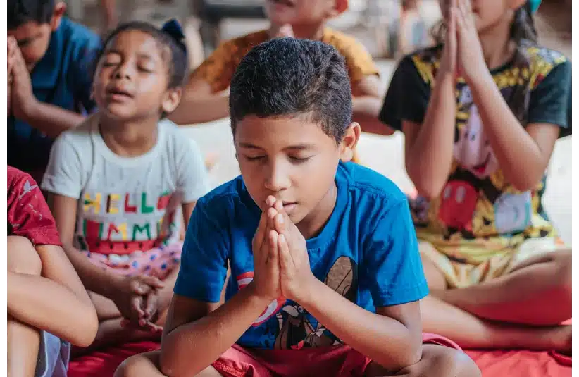 A group of kids, children, eyes closed, sit cross-legged, palms clasped in prayer. The atmosphere is calm and focused, suggesting a meditative setting.