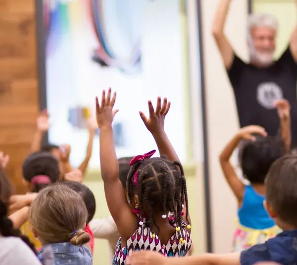 A group of young children with diverse hairstyles eagerly raise their arms in a lively children's church classroom. An adult leads them, creating a joyful, energetic atmosphere.