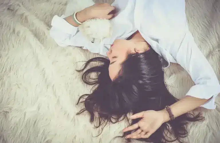 Image of Woman with long dark hair lies relaxed on a fluffy rug, holding a small white puppy. She wears a white shirt, conveying a peaceful, cozy atmosphere dreaming about old friends