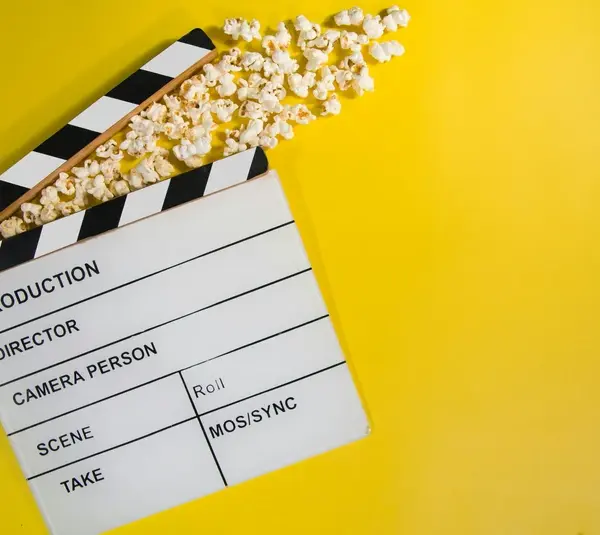 New movie releases in cinemas and theaters this weekend are shown by a film clapperboard with popcorn spilling out, lying on a vibrant yellow background. The open clapper gives a playful feel, symbolizing cinema and entertainment.