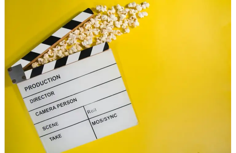 New movie releases in cinemas and theaters this weekend are shown by a film clapperboard with popcorn spilling out, lying on a vibrant yellow background. The open clapper gives a playful feel, symbolizing cinema and entertainment.