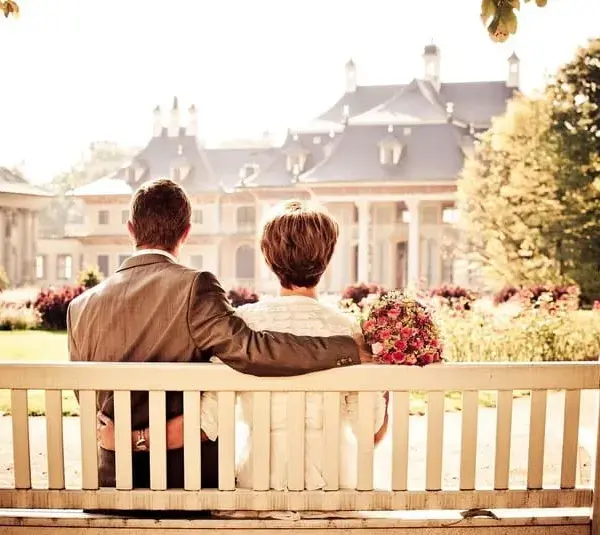 Image of a couple in wedding attire sits on Valentine's Day embracing and facing a grand resort. The scene is bathed in warm sunlight, evoking romance and serenity.