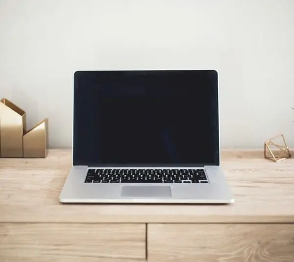 A sleek laptop doing processes with a blank screen sits on a wooden desk. To the left are gold geometric bookends, and to the right is a small potted plant, creating a minimalist and tidy workspace.