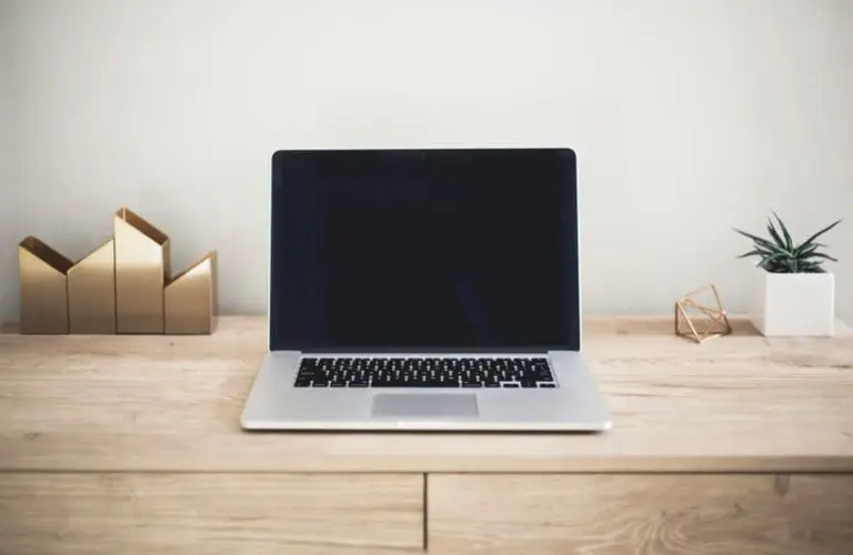 A sleek laptop doing processes with a blank screen sits on a wooden desk. To the left are gold geometric bookends, and to the right is a small potted plant, creating a minimalist and tidy workspace.