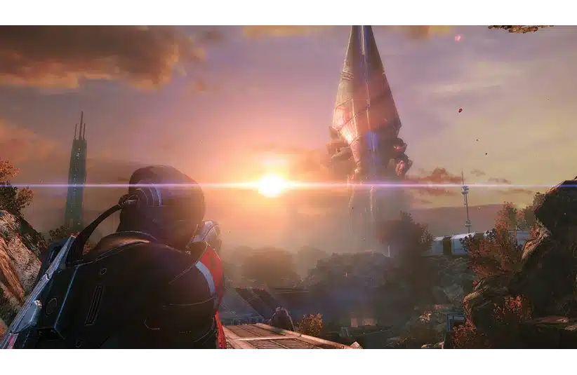 A futuristic soldier in Mass Effect in armor gazes at a towering alien structure. The setting sun casts a dramatic glow, highlighting a landscape of rocky terrain and buildings.