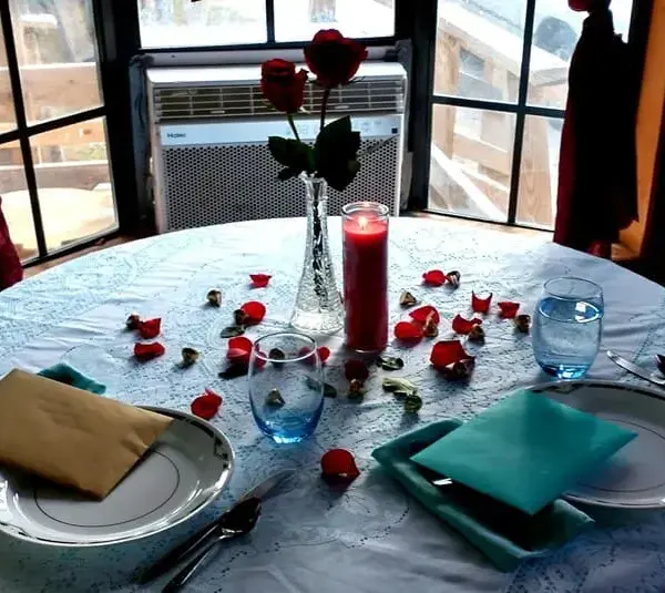 image of Romantic table setting by a window, featuring a round table with a white lace cloth, red rose petals, a candle, two plates, glasses, and a vase with roses for Valentine dinners