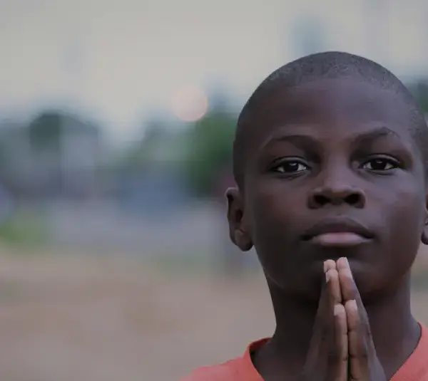 A young boy in an orange shirt stands outdoors with hands pressed together in a prayer-like gesture. The background is blurred, giving a serene atmosphere. Lent