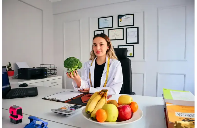 Dietician sitting behind a white desk and holding a large broccoli, clinicians guiding about viral diets