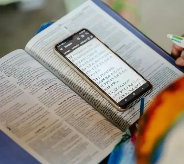 An open Bible, book, with a smartphone on top displaying text, held by a person with a pen in hand, indicating study or research, at church, home, or coffee shop. Bright and focused tone.