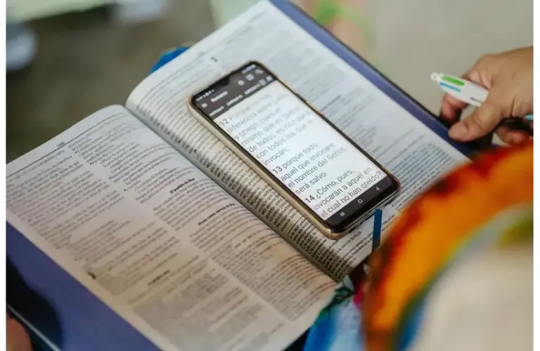 An open Bible, book, with a smartphone on top displaying text, held by a person with a pen in hand, indicating study or research, at church, home, or coffee shop. Bright and focused tone.